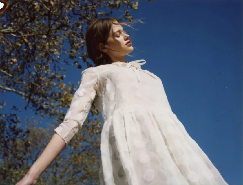 Young woman in a white dress with polka dots stands with arms outstretched, gazing upward into the bright blue sky with dappled sunlight.