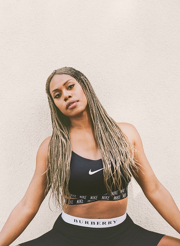 Athletic woman posing in sports bra and athletic shorts against a white wall. She is wearing braids and looking at the camera.