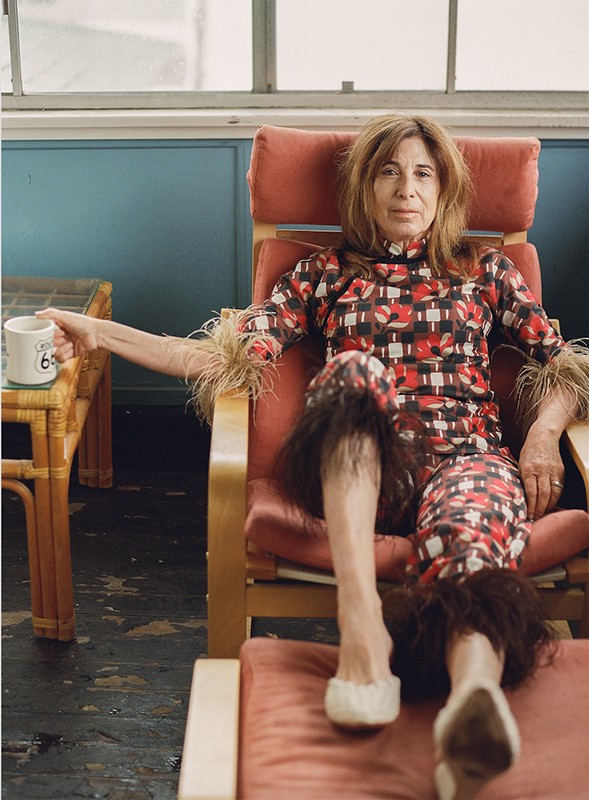 Adult woman sitting in a retro chair in patterned top and pants with feathered trim, holding a mug in an indoor setting.