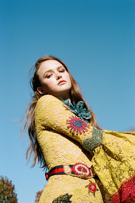 Young woman in a yellow lace dress with floral embellishments poses against a clear blue sky, viewed from a low angle.