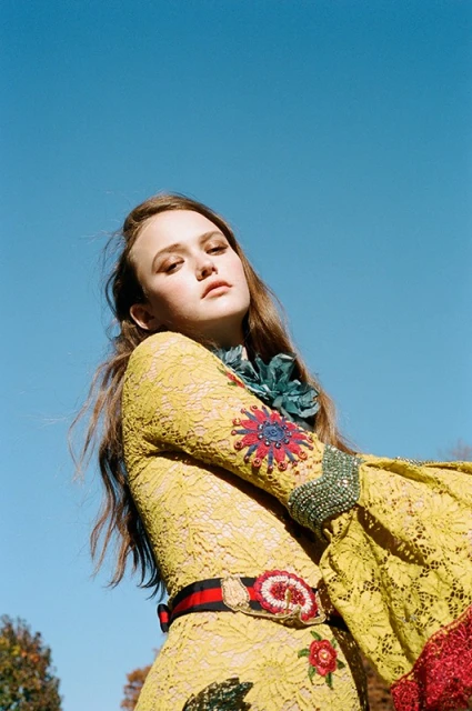 Young woman in a yellow lace dress with floral embellishments poses against a clear blue sky, viewed from a low angle.