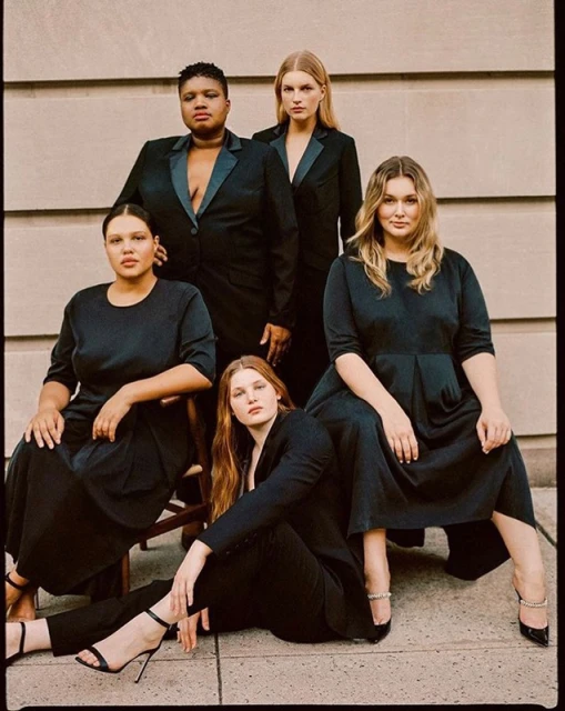 Five diverse women in formal black attire pose outdoors against a neutral wall. Models wear dresses, pantsuits, and heels in a portrait.