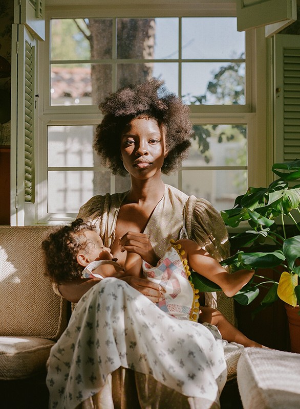 An African-American woman sits inside by a window and holds a baby in her arms while breastfeeding. Green potted plants nearby.