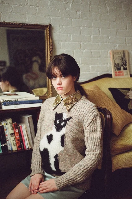 Portrait of a young woman in a cat sweater sitting on a yellow couch in front of a white brick wall and bookshelf.