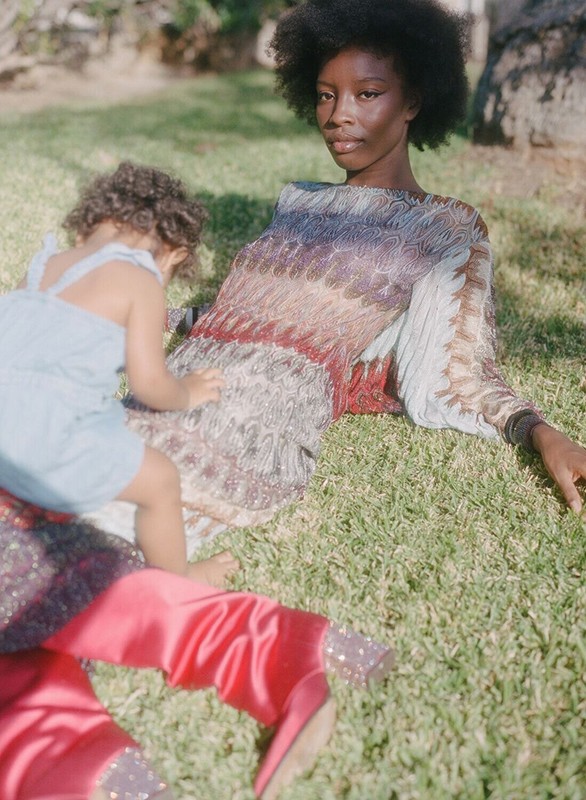 Mother and child in a grassy outdoor setting. The woman with afro hair is reclined while the toddler plays beside her.