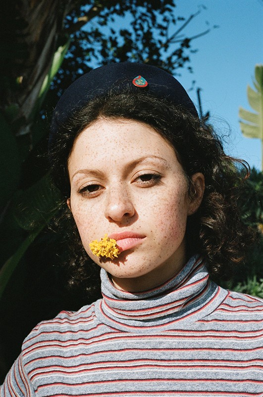 Woman with curly hair wears a black hat and a striped turtleneck. A yellow flower is held between her lip and teeth.