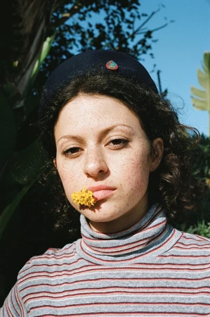Woman with curly hair wears a black hat and a striped turtleneck. A yellow flower is held between her lip and teeth.