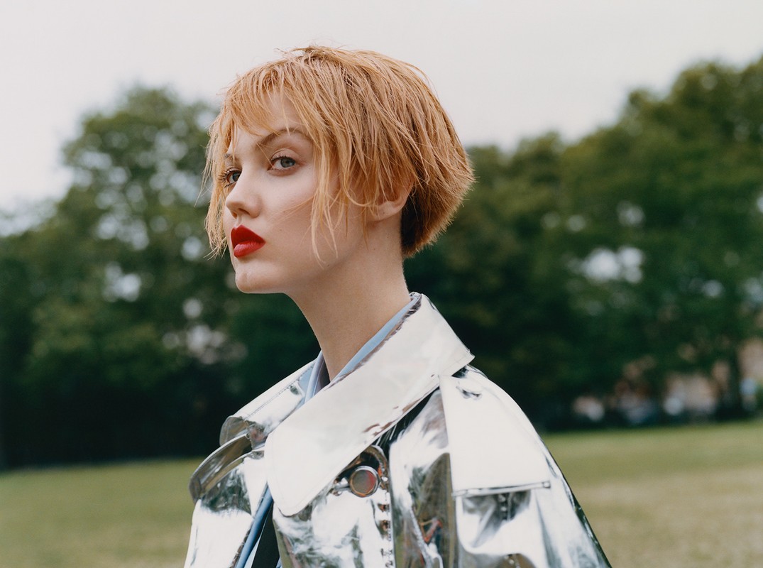 A young woman with short, light-brown hair and red lipstick wearing a silver jacket poses outdoors against a green lawn and trees.