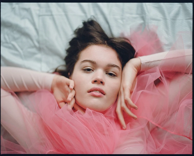 Portrait of a young woman lying on a light blue bed covered in pink tulle fabric, looking at the camera with a neutral expression.