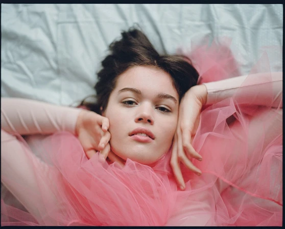 Portrait of a young woman lying on a light blue bed covered in pink tulle fabric, looking at the camera with a neutral expression.