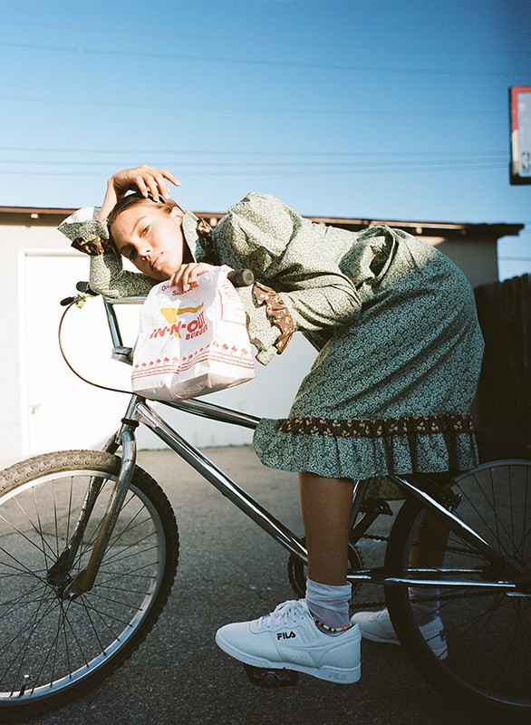 Woman in floral dress posing with In-N-Out Burger bag on bicycle in urban setting with building and bright sky.