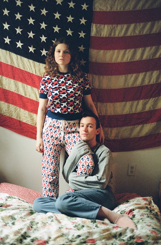 A young couple poses in front of a vintage American flag, wearing matching airplane-patterned pajamas on a floral patterned bedspread.