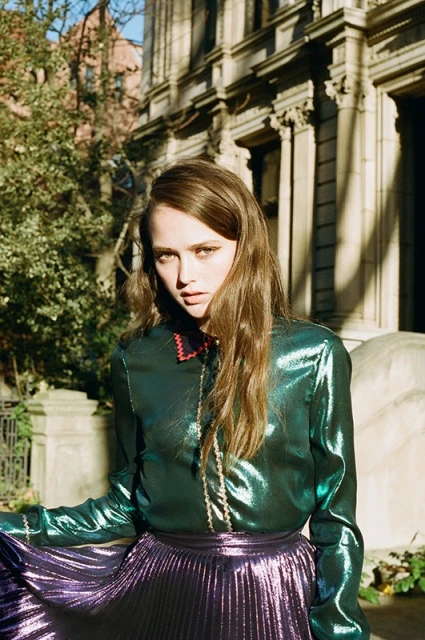 A stylish woman with long brown hair poses outdoors in a shimmering green top and pleated purple skirt, with a building behind her.