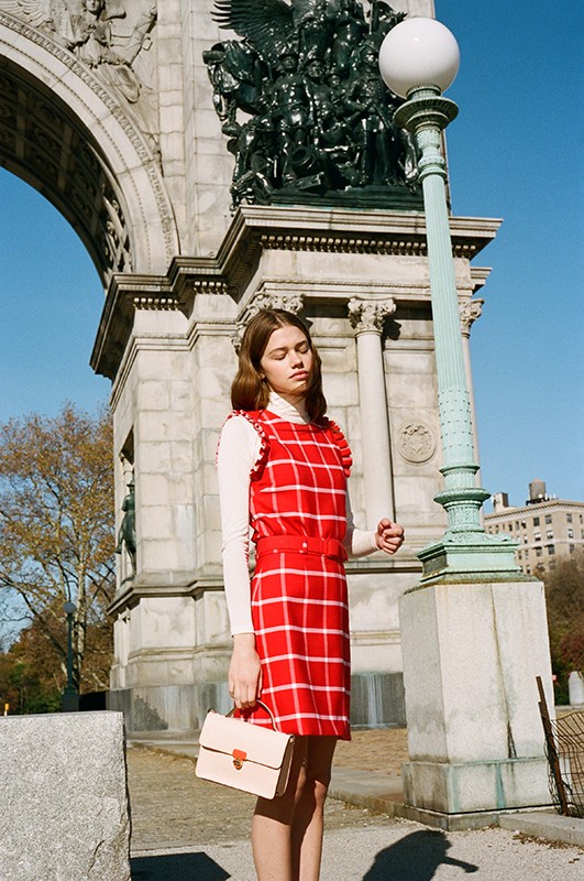Young woman poses in a red plaid dress with a cream-colored purse in an urban outdoor setting with monument arch.