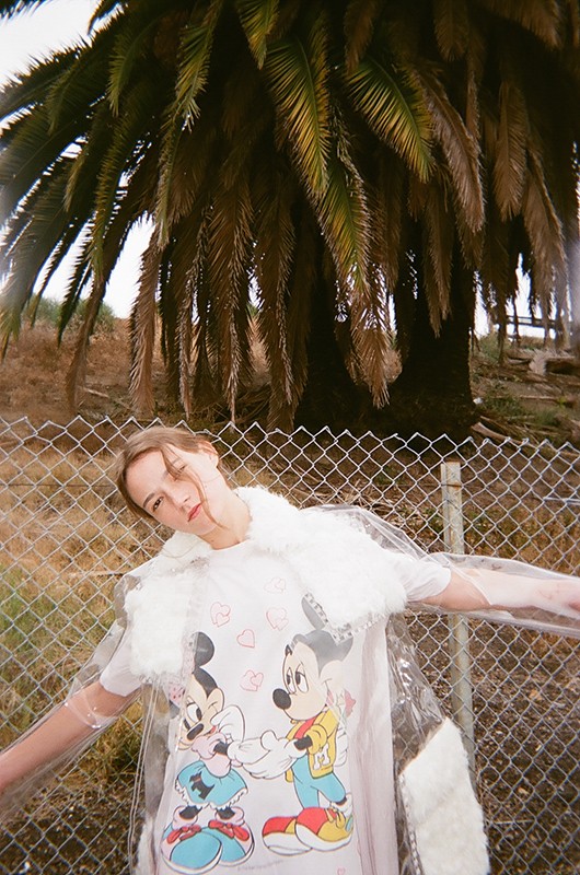Adult woman in a transparent coat with fur trim poses against a chain link fence with palm trees in the background on a sunny day.