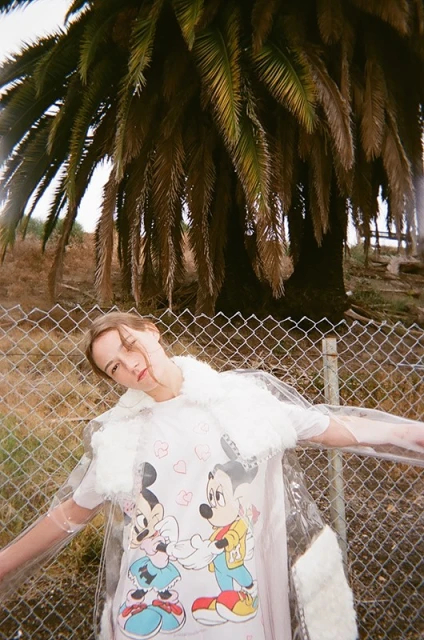Adult woman in a transparent coat with fur trim poses against a chain link fence with palm trees in the background on a sunny day.
