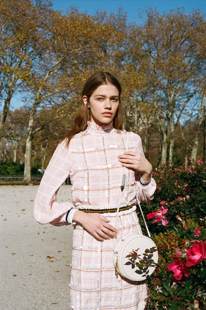 Fashionable woman in a pink dress poses in a park with blooming roses, trees, and a blue sky in the background, showcasing outdoor style.