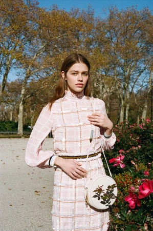 Fashionable woman in a pink dress poses in a park with blooming roses, trees, and a blue sky in the background, showcasing outdoor style.