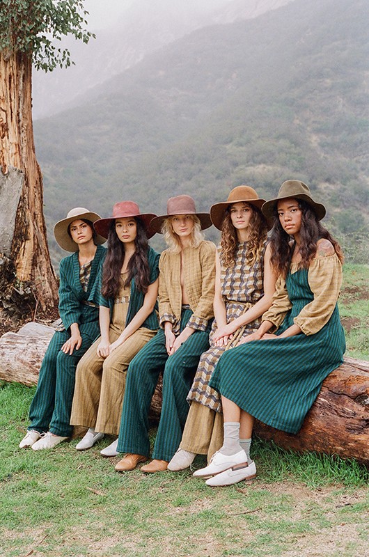 Five women wearing hats and coordinated outfits sit on a fallen tree in a misty, mountainous outdoor setting with tall trees in the background.