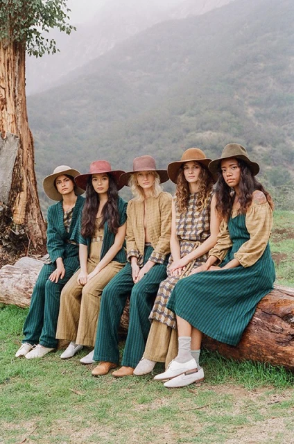 Five women wearing hats and coordinated outfits sit on a fallen tree in a misty, mountainous outdoor setting with tall trees in the background.