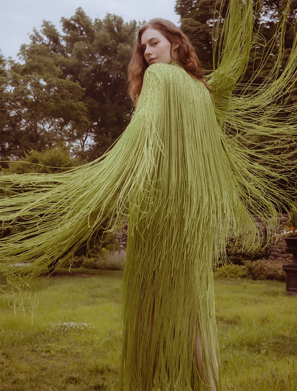 Adult woman in long, light-green, shredded fabric dress posing in a lush grassy outdoor setting with trees in the background.