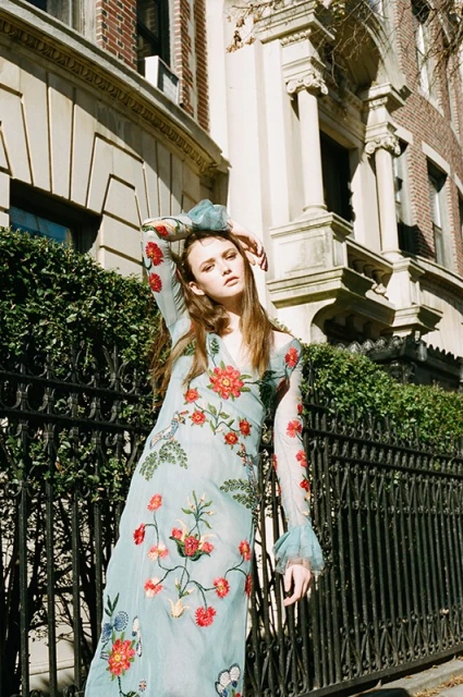 Woman in a floral dress poses in front of a building with a black metal fence; outdoor fashion shot with natural light.