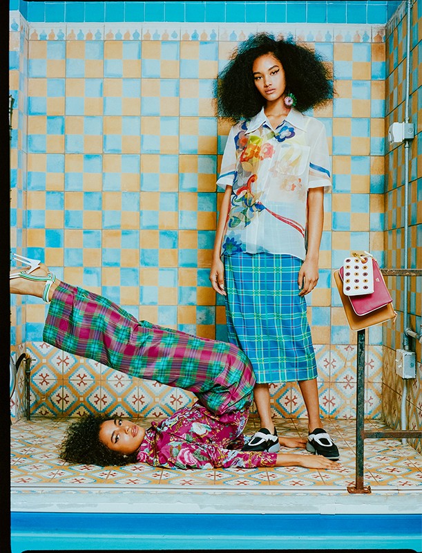Two young women in a tiled bathroom: one lying on the floor with legs up, wearing a floral top and plaid pants; one standing in a skirt.