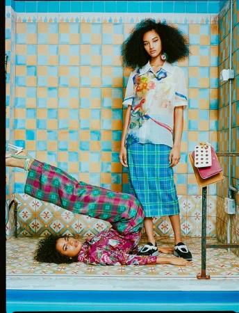 Two young women in a tiled bathroom: one lying on the floor with legs up, wearing a floral top and plaid pants; one standing in a skirt.