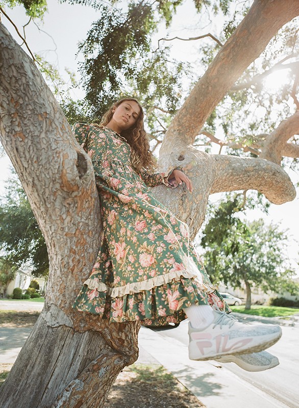 A young woman in a floral dress sits in a tree, wearing sneakers. Sunlight illuminates her and the tree branches overhead.