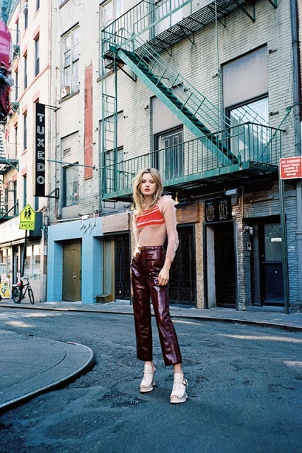 A woman in shiny maroon pants and a striped crop top poses on a city street with buildings and a fire escape in the background.
