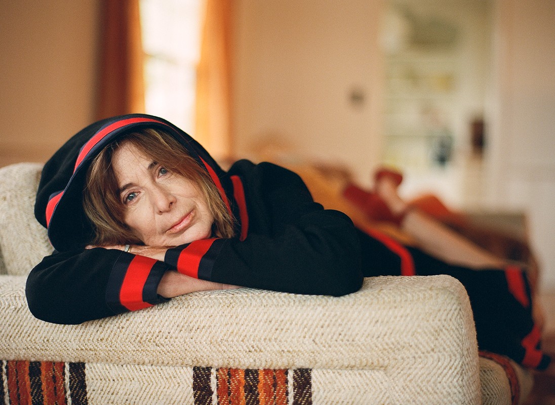 An elderly woman reclines on a textured sofa in a relaxed pose, wearing a black hooded sweatshirt with red stripes in a warm, indoor setting.