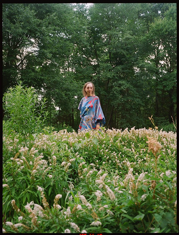 An adult female standing in a field of white flowers with trees visible in the background, captured in an eye-level outdoor shot.