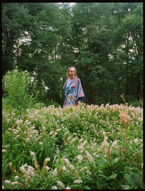 An adult female standing in a field of white flowers with trees visible in the background, captured in an eye-level outdoor shot.