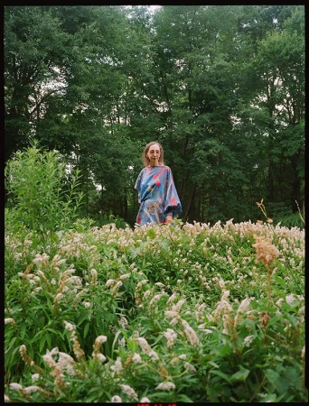 An adult female standing in a field of white flowers with trees visible in the background, captured in an eye-level outdoor shot.
