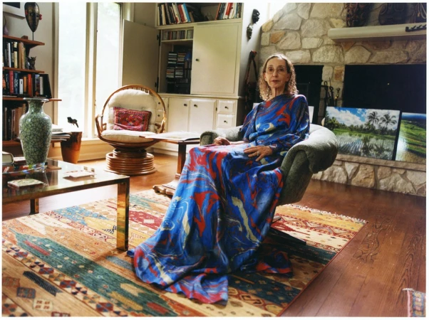 Older woman in colorful patterned dress seated in armchair in a living room with stone fireplace, bookshelves, and paintings.