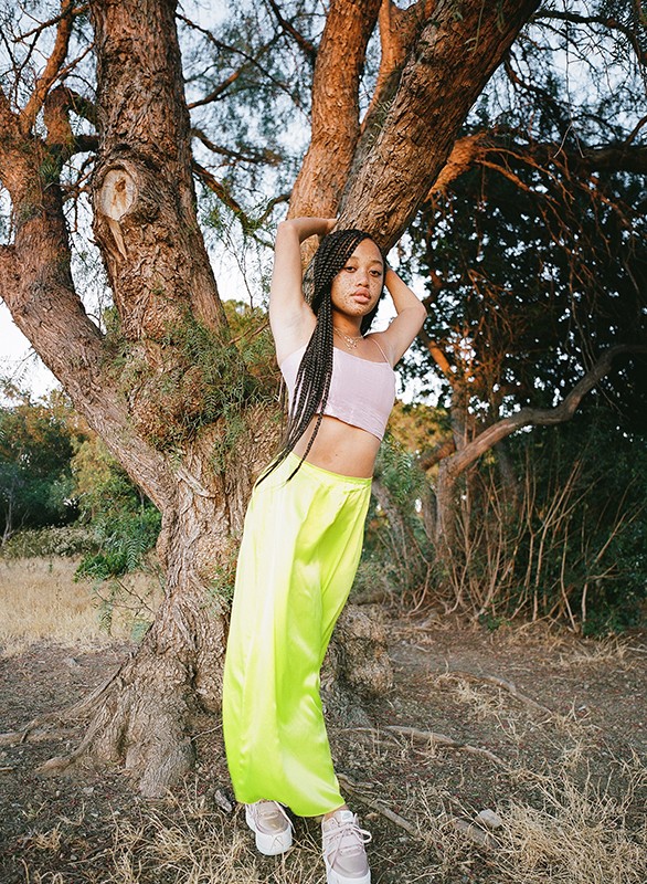 Young adult woman in neon green pants and lavender top leans against a tree in an outdoor setting with soft, natural lighting.