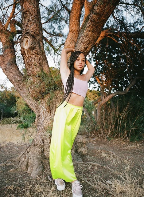 Young adult woman in neon green pants and lavender top leans against a tree in an outdoor setting with soft, natural lighting.