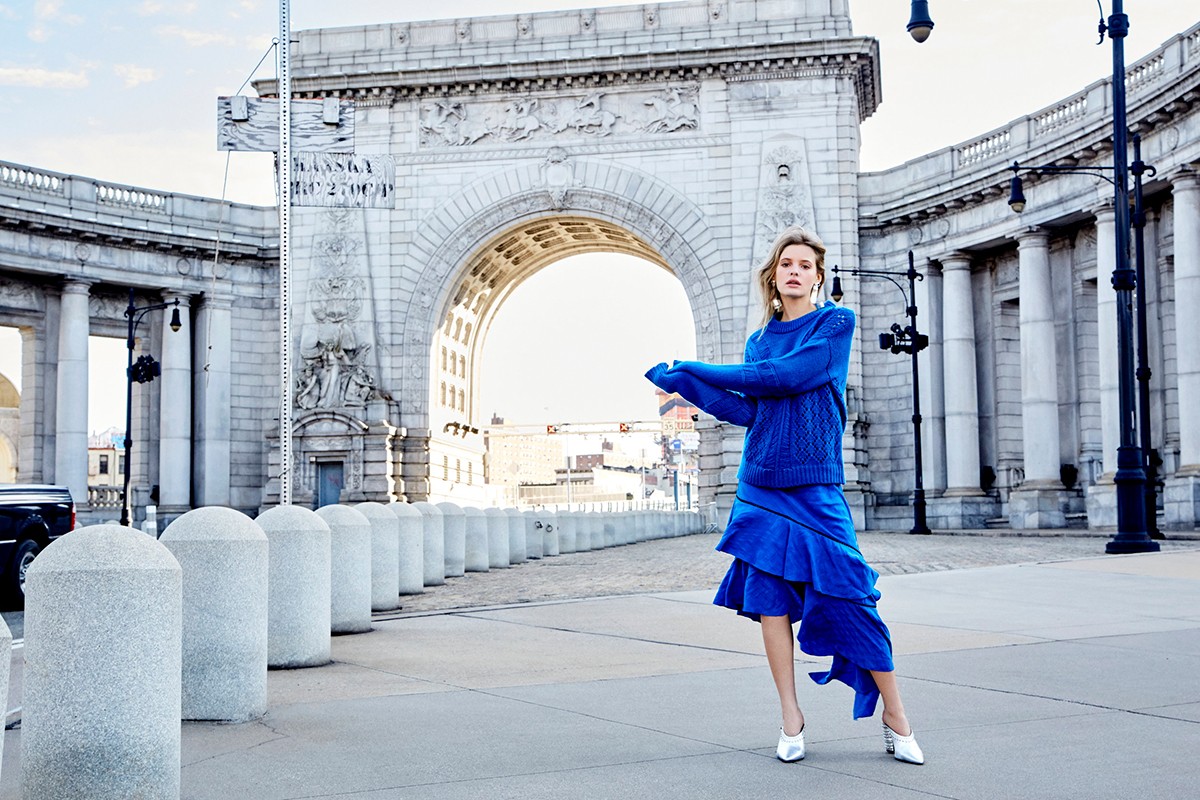 Fashion shot of a blonde woman in a bright blue outfit on a pedestrian walkway in front of Manhattan Bridge against an outdoor backdrop.