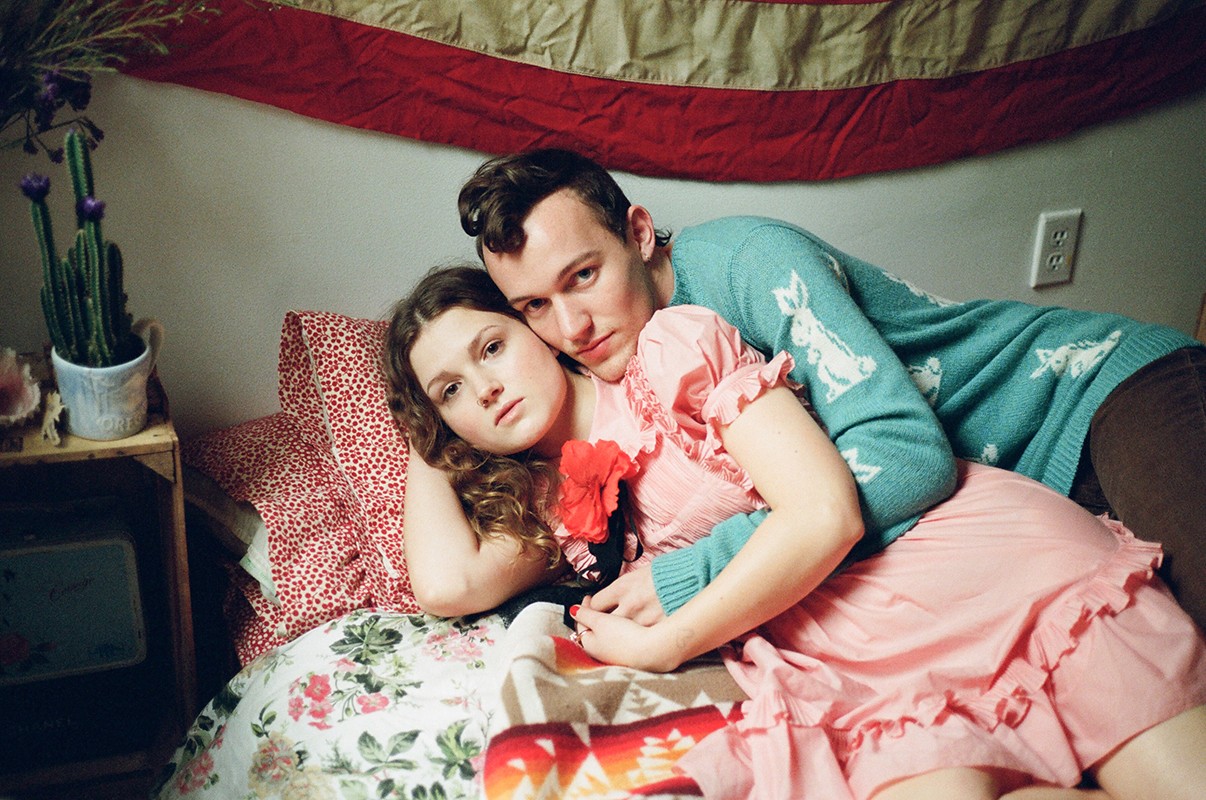 Couple embraces on a floral bed under an American flag; the woman in a pink dress and the man in a bunny sweater.