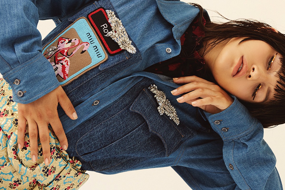 A stylish young woman with gold eyeliner poses wearing a denim jacket with embellished patches and a printed skirt against a plain background.