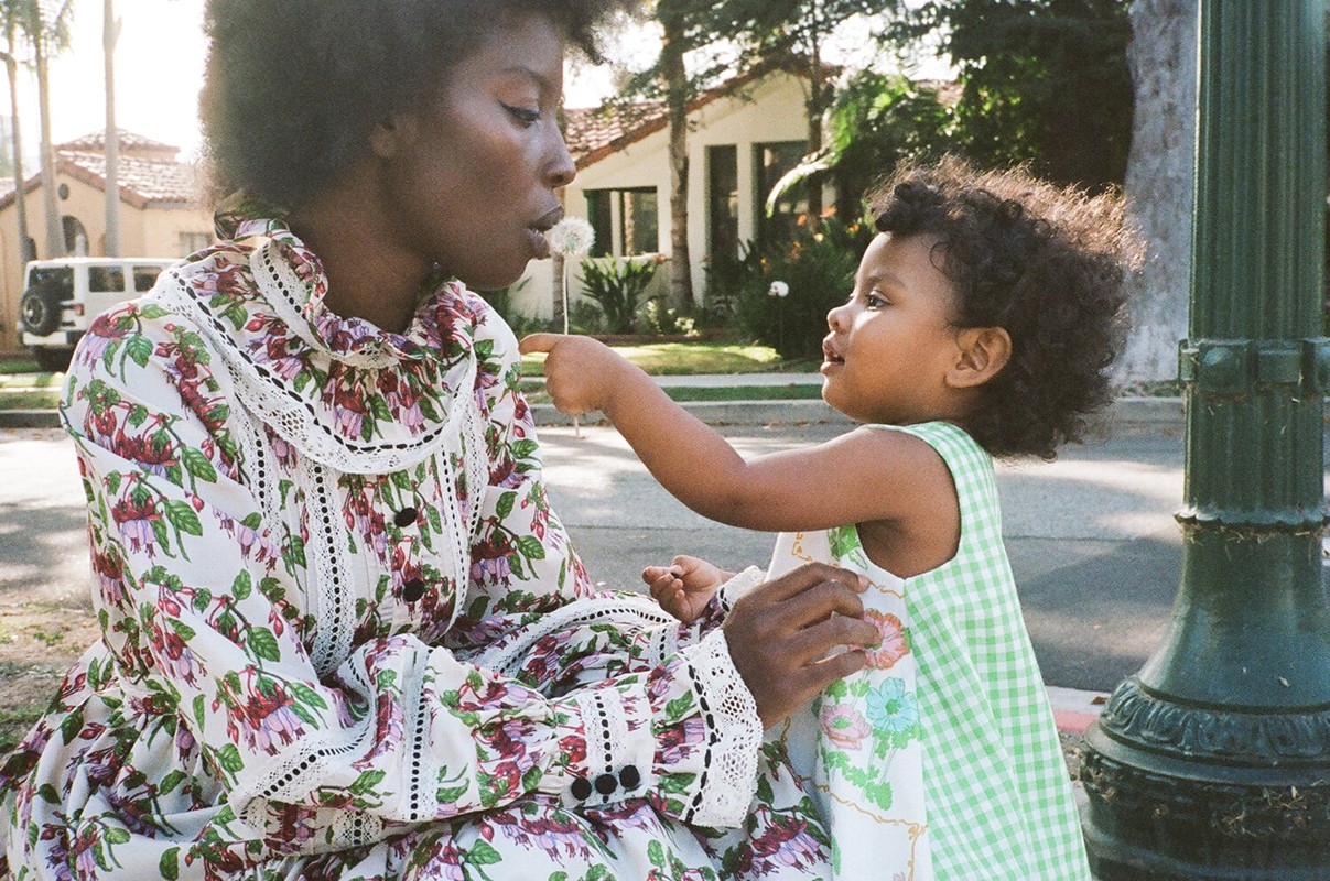 African-American mother and child share a tender moment in a sunlit outdoor setting, with the child playfully reaching towards a dandelion.