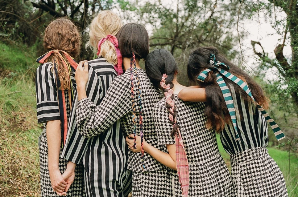 Group of five young women with colorful ribbons in their hair stand closely together in an outdoor setting, showcasing fashion and friendship.