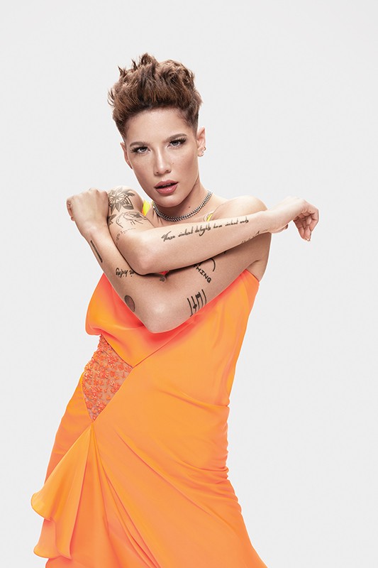 A person with short brown hair, tattoos, and a bright orange dress stands in a studio setting posing with their arms crossed.