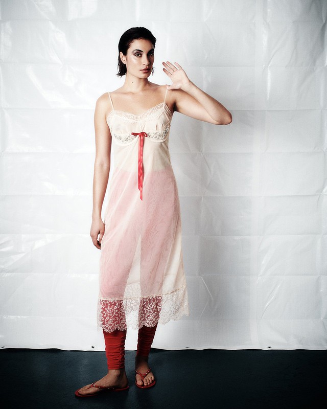 Woman in slip dress posing indoors against a white background. She wears red leggings and sandals, and has dark short hair.
