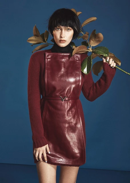 Fashion shot of a woman with dark hair wearing a short, red leather dress over a long-sleeved shirt, and holding a leafy branch.