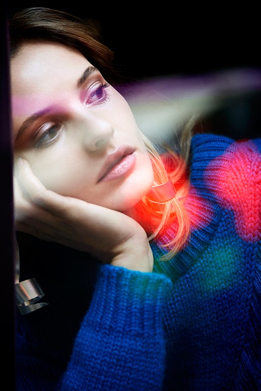 Portrait of a woman with shoulder-length brown hair, resting her chin on her hand and wearing a blue sweater with colorful accents.
