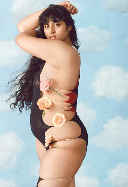 Model poses in a black bodysuit with flowers and beaded strands against a light blue cloudy background in a beauty fashion editorial.