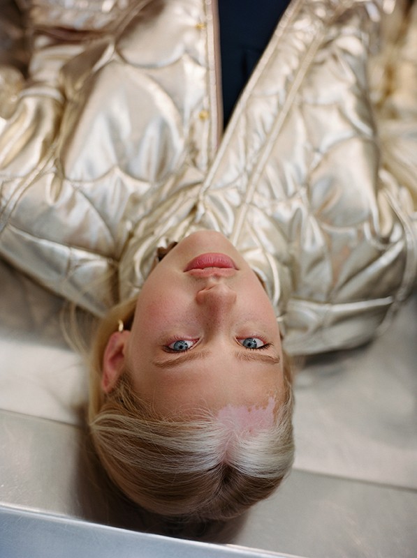 Close-up portrait of a blonde woman wearing a metallic gold quilted puffer jacket, shot from an overhead perspective.
