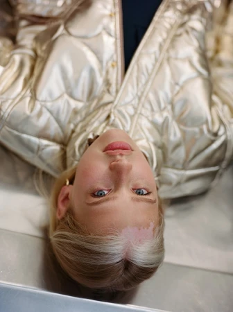 Close-up portrait of a blonde woman wearing a metallic gold quilted puffer jacket, shot from an overhead perspective.