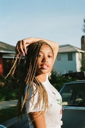 Young Black woman with long braided blonde hair holding her hand to her head, wearing a white t-shirt outdoors in Los Angeles.